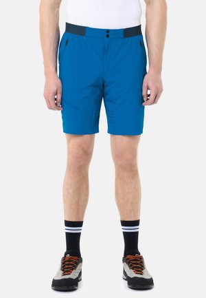 Uomo che indossa pantaloncini blu con cerniera, calzini neri con strisce bianche e scarpe da trekking grigie con lacci arancioni, in piedi contro uno sfondo bianco.