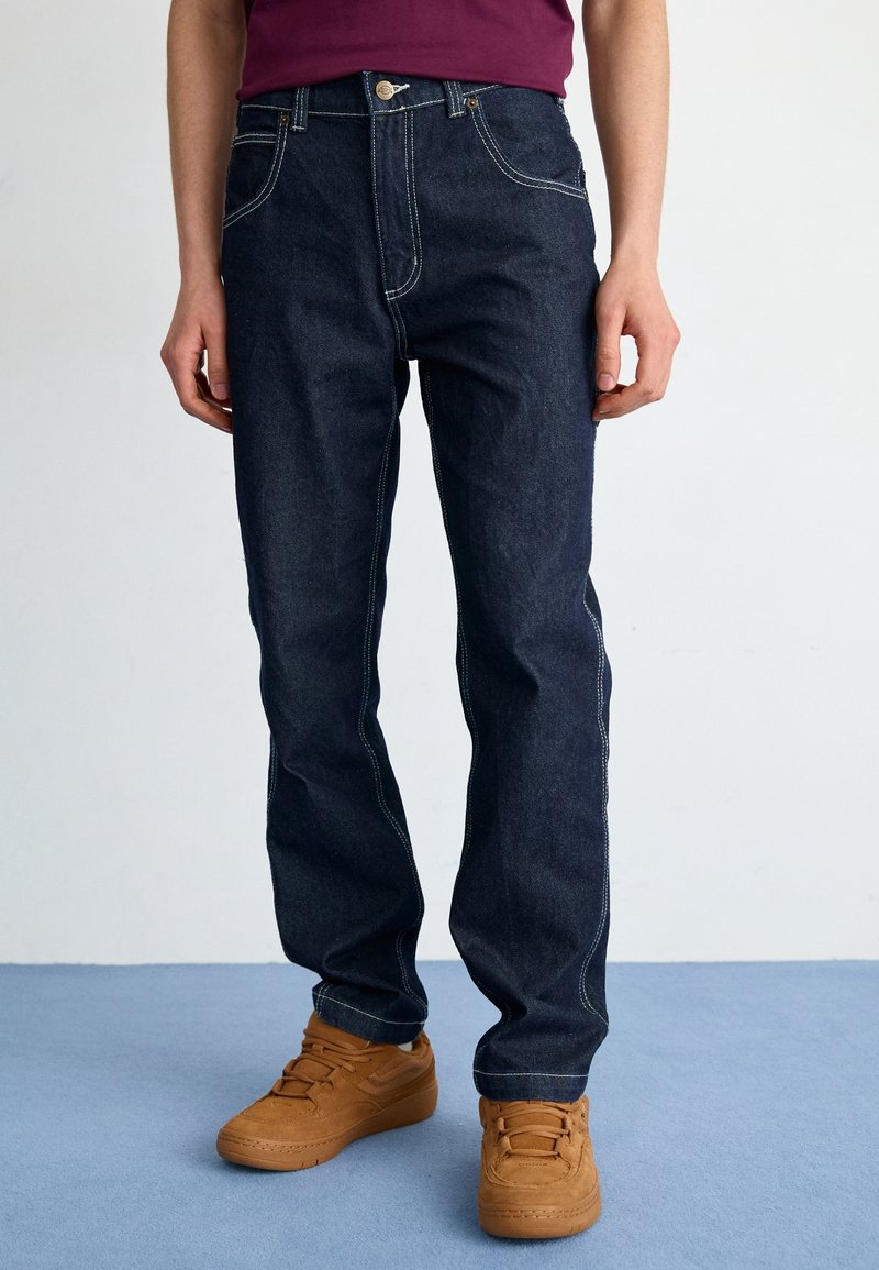 Homme portant un jean bleu foncé avec des coutures blanches et des baskets marron, debout sur un sol bleu devant un mur blanc.