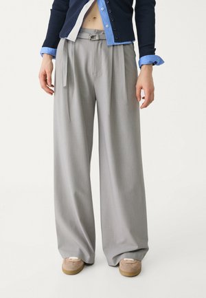 Persoon draagt hooggetailleerde, wijde grijze broek met riem, marineblauwe cardigan met knopen, blauwe overhemdmanchetten en beige sneakers.