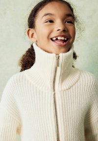 Pull en tricot côtelé blanc avec col montant et demi-zip. Présente une texture douce et un design décontracté, parfait pour le superposition.