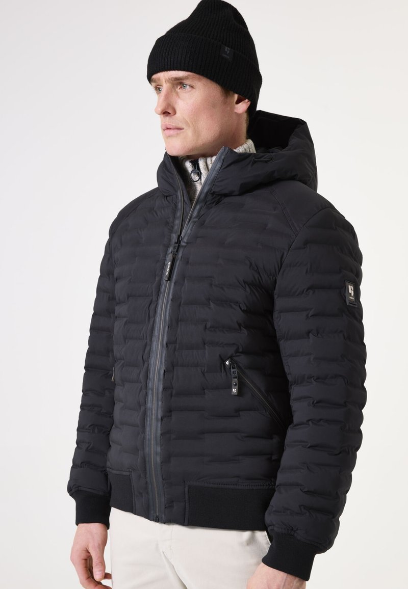 Schwarze gepolsterte Jacke mit Kapuze, Reißverschluss, seitlichen Taschen, gerippten Bündchen und glatter Textur. Getragen über einem leichten Pullover.