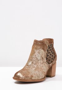 Brązowe botki sięgające kostki z teksturowanym, błyszczącym wykończeniem, otwartymi panelami z siatki i masywnym drewnianym obcasem. Okrąły czubek i elastyczne wstawki po bokach.