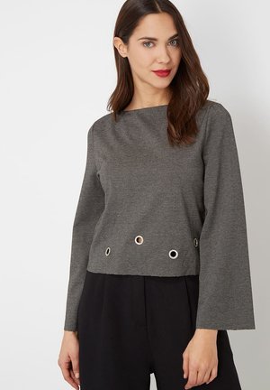 Mujer con cabello castaño que lleva una camiseta gris de manga larga con ojales de metal cerca del dobladillo y pantalones negros, de pie contra un fondo blanco.