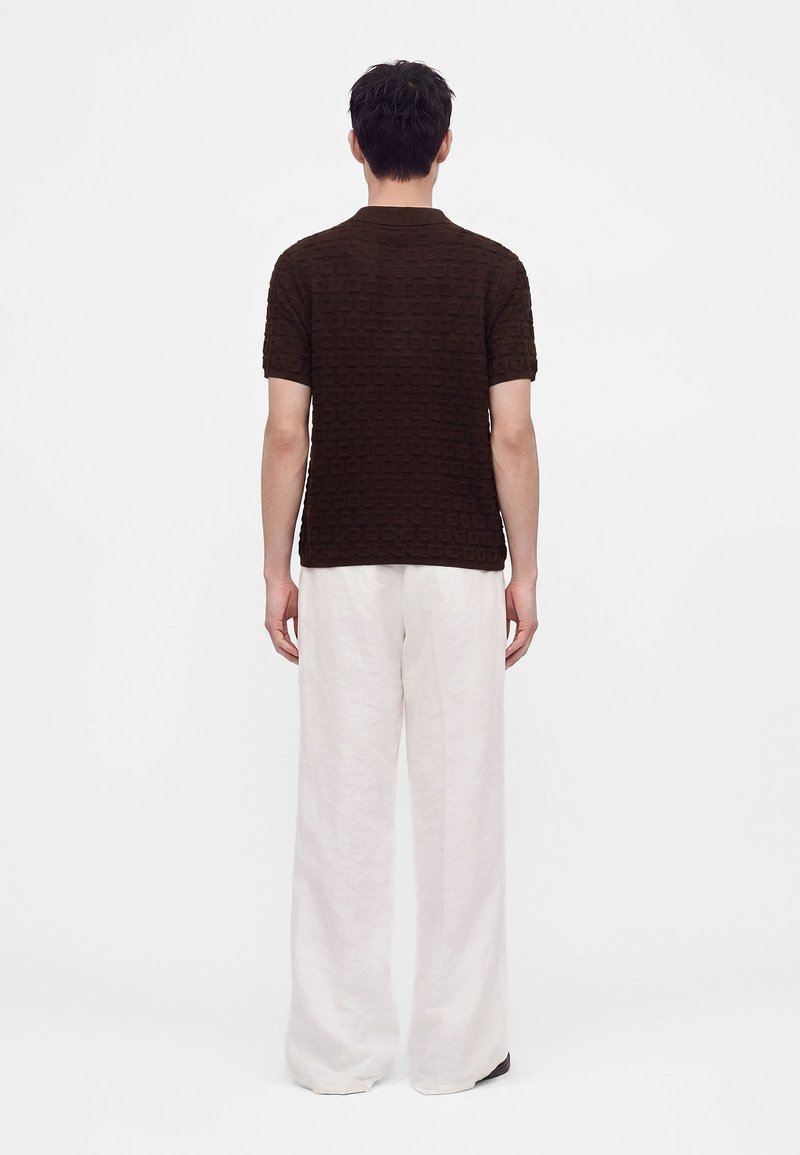 Persona de pie de espaldas, vistiendo una camisa de manga corta texturizada color marrón oscuro y pantalones blancos holgados contra un fondo blanco liso.