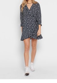 Navyblaues Blumenkleid mit langen Ärmeln, einem V-Ausschnitt und einem Rüschensaum. Der Stoff ist mit kleinen weißen Blumen versehen und hat eine weiche Textur. Trägt es mit weißen Sneakers.