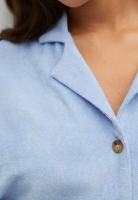 Detailní záběr na osobu, která má na sobě světle modrou košili s límcem a hnědým knoflíkem zapnutým poblíž hrudi.