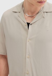 Camisa de manga corta, de un suave color beige claro, hecha de una tela lisa con cierre de botones en la parte delantera, cuello con muescas y un sutil logo bordado en el pecho.
