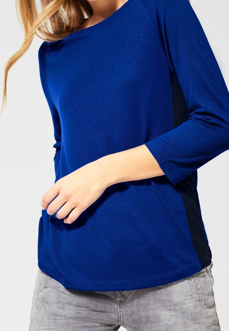 Haut à manches longues bleu avec un col rond et des accents de panneau noir sur les côtés, fabriqué à partir d'un tissu lisse et léger. Stylé avec une coupe décontractée.