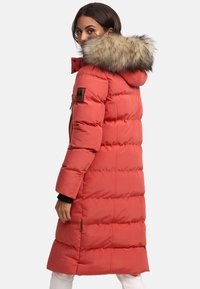 Geacă roșie pufoasă cu glugă căptușită cu blană, având un design quilted, fermoare pe laterale și o etichetă cu logo pe mâneca stângă.