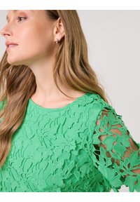 Grünes Spitzenoberteil mit floralem Muster und offenen Ärmeln. Der Stoff hat ein strukturiertes, geschichtetes Aussehen und einen runden Ausschnitt.