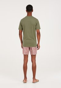 Camiseta de algodón verde oliva de manga corta y corte relajado, combinada con bañador rojo con estampado y sandalias, vista desde la parte de atrás.