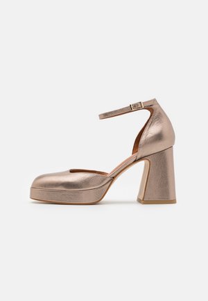 Chaussure à talon haut couleur or rose avec bout rond, bride de cheville avec boucle, semelle plateforme et talon bloc sur fond clair uni.