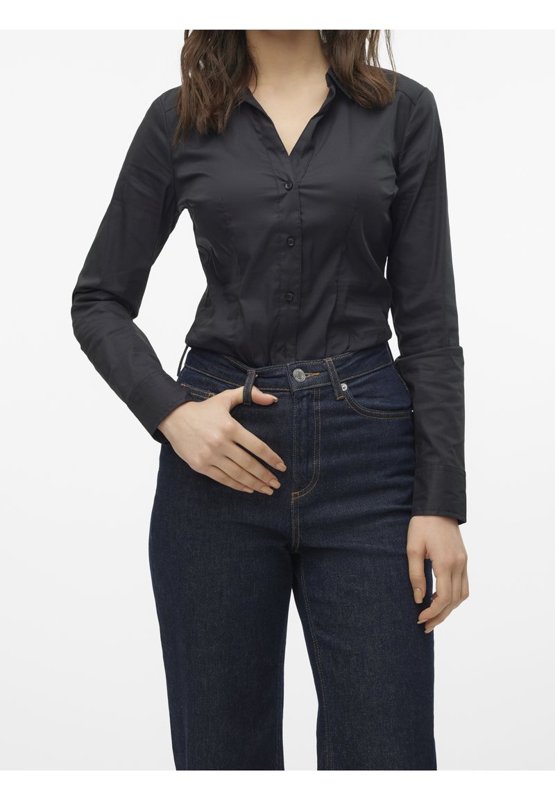 Femme portant une chemise noire cintrée boutonnée rentrée dans un jean taille haute bleu foncé, debout avec une main près de la ceinture.