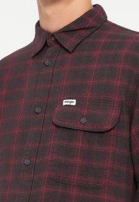 Camicia di flanella a quadri rossi e neri con colletto button-up, taschino e etichetta con marchio. Presenta un tessuto testurizzato e bottoni scuri.