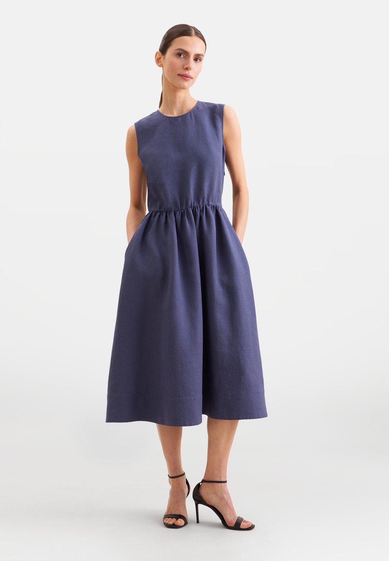 Femme debout, les mains dans les poches, portant une robe midi sans manches bleu marine et des sandales noires à talons hauts sur un fond uni clair.