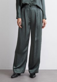 Pantalons larges taille haute en vert foncé, en tissu brillant. Comprend une fermeture à bouton, des plis et une texture lisse.