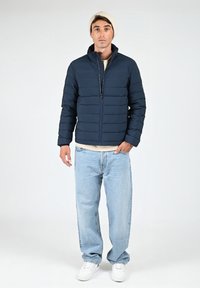 Veste matelassée bleue avec un motif en quilt horizontal, col haut et fermeture éclair à l'avant. Associée à un jean bleu clair et des baskets blanches.