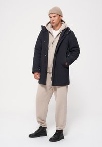 Granatowy długi płaszcz, z zamkiem z przodu, kapturem i kieszeniami. Beżowa bluza z kapturem i spodnie dresowe pod spodniami. Czarne botki za kostkę. Jasnobeżowa dziergana czapka.