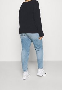 Henkilö seisoo selin, päällä musta neule, vaaleansiniset farkut, valkoiset sukat ja valkoiset lenkkarit yhtenäisellä vaalealla taustalla.