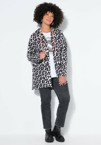 Lang jakke med leopardprint og orange snor, båret over en hvid t-shirt med grafik og sorte jeans. Sorte ankelstøvler fuldender outfittet.