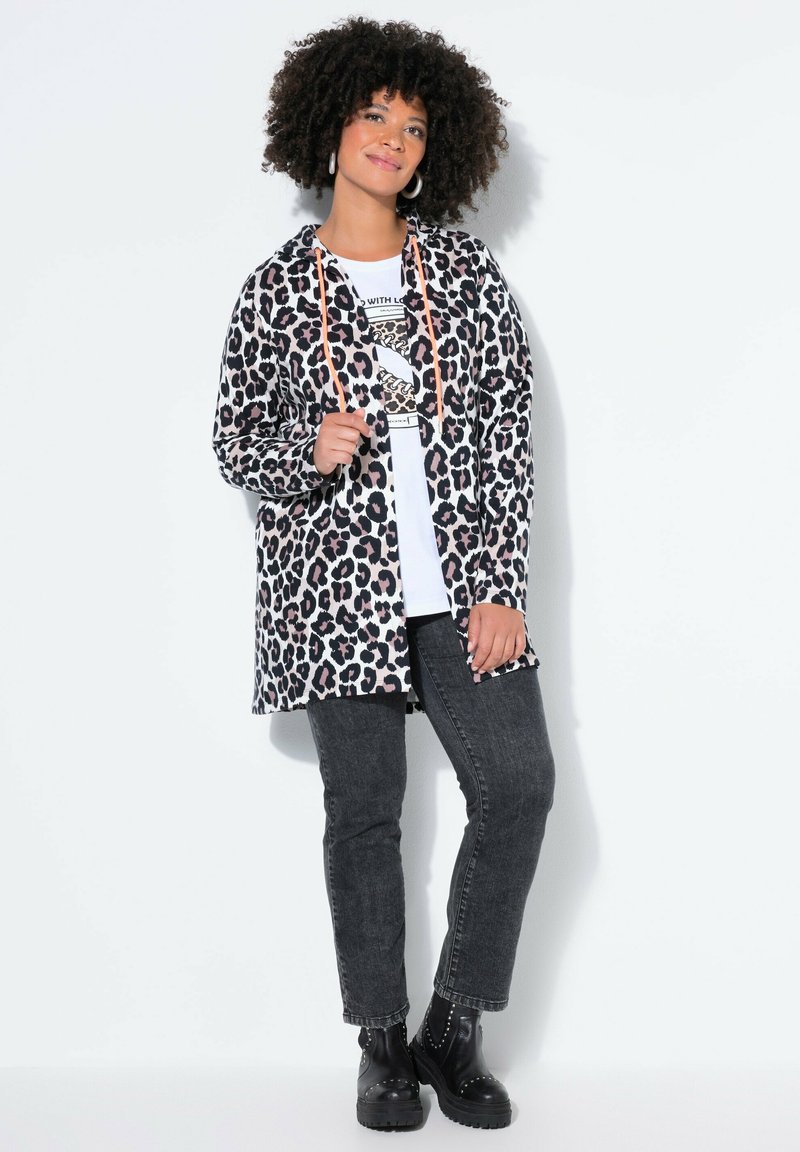 Lang jakke med leopardprint og orange snor, båret over en hvid t-shirt med grafik og sorte jeans. Sorte ankelstøvler fuldender outfittet.