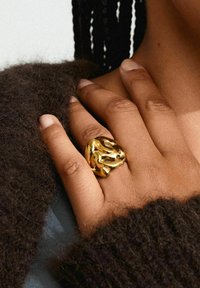 Bague sculpturale en or aux formes organiques portée sur une main, au-dessus d'un pull sombre et texturé. Le teint de la peau et la couleur des ongles sont neutres.