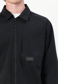 Chaqueta negra con cremallera hecha de una tela suave, con un cuello, dos bolsillos frontales y una etiqueta de marca en el bolsillo izquierdo.