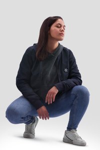 Lässiges Outfit mit einer dunklen Bomberjacke, grauem Hoodie, blauen Jeans und grauen Sneakers. Die Jacke hat gerippte Bündchen und ein gesticktes Logo.