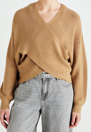 Femme portant un pull enveloppant en maille côtelée couleur chameau et un jean taille haute gris clair avec une ceinture à double boucle.