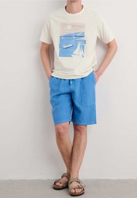 Camiseta ligera beige para hombre con un gráfico azul de barcos y un sol. Combinada con pantalones cortos de lino azul y sandalias marrones.