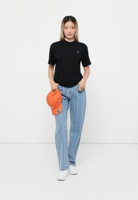 Black short-sleeve t-shirt, light blue striped jeans, orange cap in hand, white sneakers, model standing against a plain background.