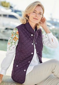 Lila gestepptes Weste mit weißen, floral gemusterten Ärmeln, ausgestattet mit Druckknöpfen. Kombiniert mit einem weißen Shirt und hellbeigen Hosen.