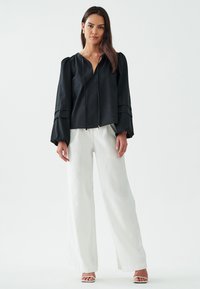 Blusa nera a maniche lunghe con chiusura con zip e maniche a sbuffo, abbinata a pantaloni bianchi a gamba larga e sandali con cinturino.