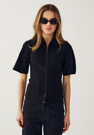 Jonge vrouw die een zwart shirt met korte mouwen en ritssluiting draagt, zwarte spijkerbroek en smalle zwarte zonnebril, staand tegen een effen lichte achtergrond.