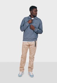 Lyseblå pullover med opretstående krave og logo, parret med beige bukser og sko med blå accenter. Glat stof og afslappet pasform.