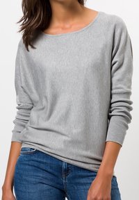 Ljust grå långärmad tröja i mjukt tyg, med en vid halsringning, ribbade ärmslut och en avslappnad passform. Kombinerad med blå jeans.