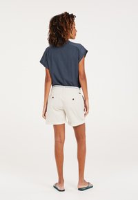 Marineblauw shirt met korte mouwen in combinatie met lichtbeige shorts met een omgeslagen zoom. De look wordt aangevuld met casual slippers.