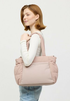 Jonge vrouw met rood haar, witte lange mouwen en een blauwe spijkerbroek, draagt een grote lichtroze gewatteerde draagtas over haar schouder.