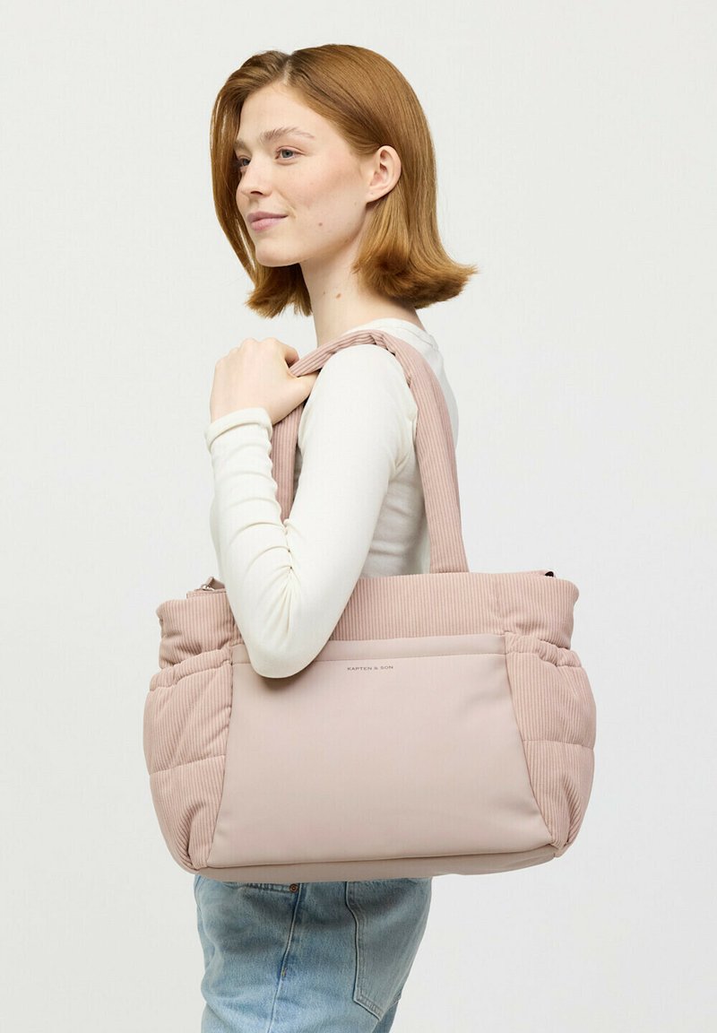 Młoda kobieta z czerwonymi włosami, ubrana w białą bluzkę z długim rękawem i niebieskie dżinsy, niesie dużą, jasnoróżową, pikowaną torbę na ramię.