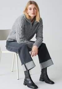Maglione grigio a coste con mezzo zip, jeans scuri a vita alta con orli arrotolati e stivali neri con tacco fino al ginocchio. Sedia minimalista sullo sfondo.