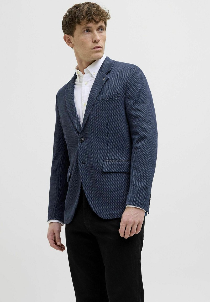 Tumes tekstuuriga bleiser ühe nupuga, klappidega krae ja kahe esisegmendiga, kombineeritud valge nööbipaelaga särk ja mustad püksid.