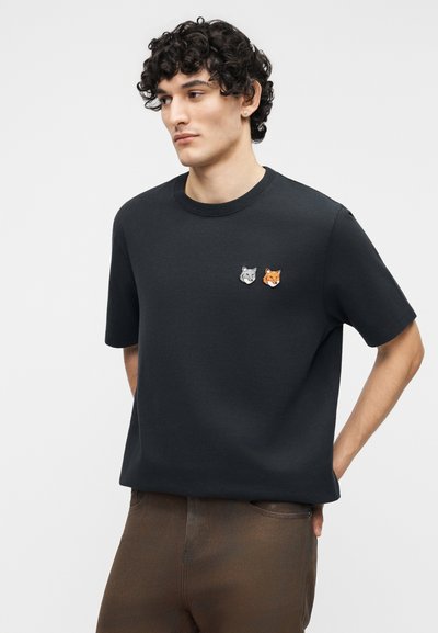 Άνδρας με σγουρά μαλλιά που φοράει μαύρο μπλουζάκι με μικρά μπαλώματα σε σχήμα κεφαλιών λύκου και αλεπούς στο στήθος, καφέ παντελόνι και τα χέρια πίσω από την πλάτη.