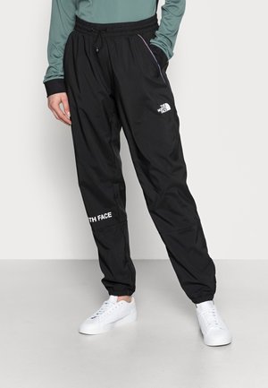 Osoba nosząca czarne spodnie dresowe The North Face z elastycznymi mankietami przy kostkach i białe sneakersy, stojąca z jedną ręką w kieszeni.