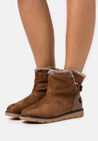 Bottines en daim marron avec bordure en fausse fourrure, bout arrondi et panneau latéral texturé. Semelle extérieure en caoutchouc plate pour traction et stabilité.