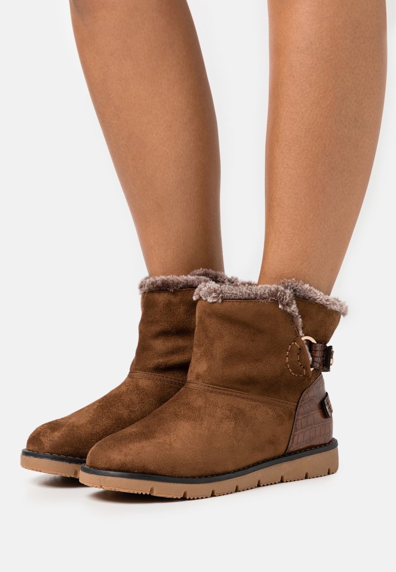 Bottines en daim marron avec bordure en fausse fourrure, bout arrondi et panneau latéral texturé. Semelle extérieure en caoutchouc plate pour traction et stabilité.