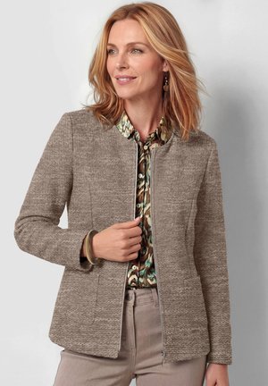 Vrouw met schouderlang blond haar draagt een getextureerd beige jasje over een groen-bruine blouse met patroon, met beige broek en een armband.