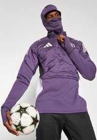 Lilla atletisk jakke med hætte, halv-zip design og logo detaljer, parret med sorte bukser. Holder en fodbold med stjernemønster.