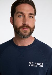 Granatowy sweatshirt z prążkowanym kołnierzem i rękawami raglanowymi. Z przodu widnieje biały napis "DREI MASTER". Miękka tekstura materiału.