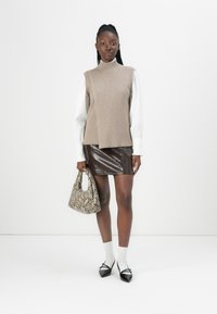 Pull sans manches beige sur une chemise blanche, associé à une jupe en cuir de serpent marron, des chaussettes blanches et des chaussures noires à bout pointu. Sac à main en imprimé serpent.
