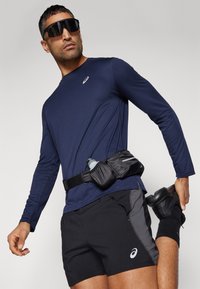 Marineblå langærmet atletisk skjorte, sorte shorts med grå detaljer, sort væskebælte, sorte sneakers og tonede solbriller.